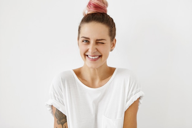 positive coquettish young European woman with pink hair knot smiling happily, blinking