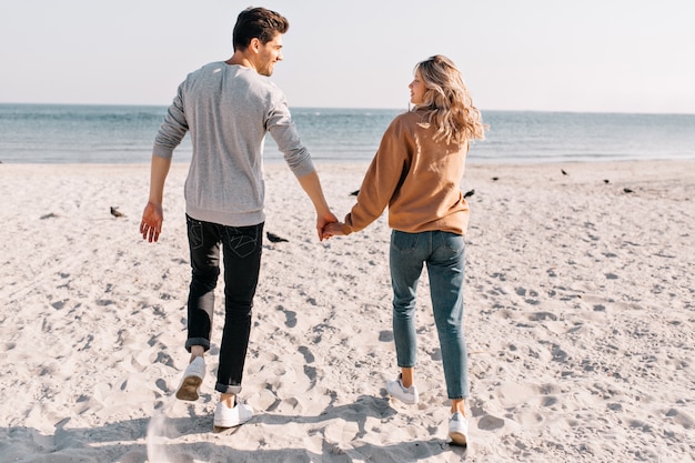笑顔で海に走るポジティブなカップル。ビーチでの休息中に彼氏と手をつないでかわいい女の子の屋外の肖像画。