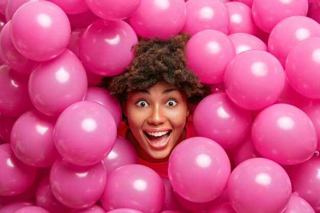positive crazy dark skinned woman looks happily, gets surprise, has fun during festive day surrounded by small inflated rosy balloons.