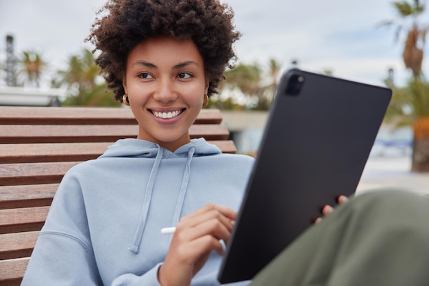 Free photo positive creative woman with curly hair dressed in casual hoodie works distantly uses touchpad and stylus for making sketches poses on wooden bench outdoors focused away modern technologies