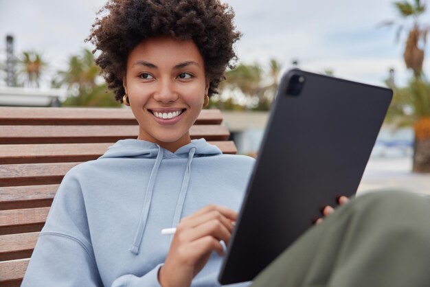 Positive creative woman with curly hair dressed in casual hoodie works distantly uses touchpad and stylus for making sketches poses on wooden bench outdoors focused away Modern technologies