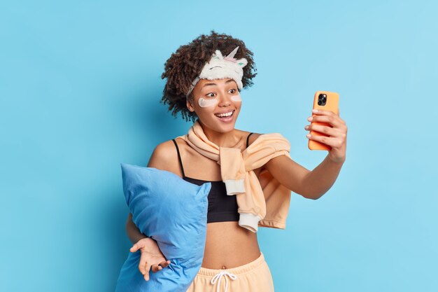 Positive culry haired young Afro American woman takes selfie via smartphone smiles happily applies collagen patches under eyes dressed in pajama holds pillow isolated over blue studio wall