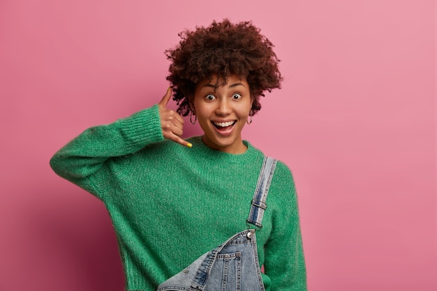 긍정적 인 곱슬 머리 아프리카 계 미국인 여성이 전화 제스처를하고, 다시 전화를 걸고, 기쁜 표정을 지으며, 녹색 스웨터를 입고, 실내에서 분홍색 벽에 포즈를 취하고, 친구들과 연락을 취합니다.