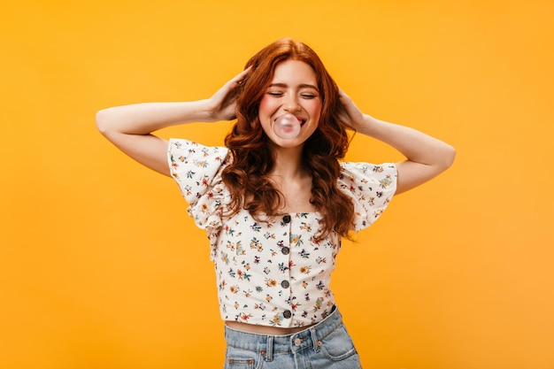 Positive curly girl in white cropped top touches her red curly hair and inflates bubble of gum