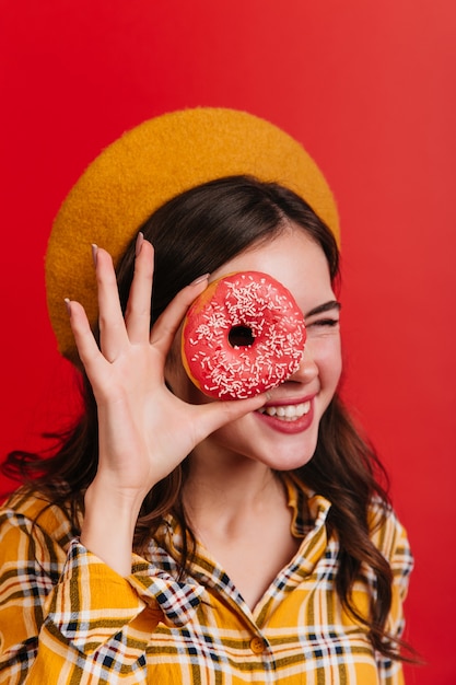 Foto gratuita la ragazza riccia positiva strizza l'occhio e si copre l'occhio con la ciambella alla fragola. donna attraente in camicia a quadri e cappello giallo in posa sul muro rosso.