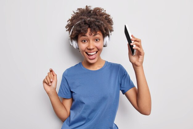 Позитивная кудрявая очаровательная женщина танцует под музыку, держит мобильный телефон, слушает любимую песню через беспроводные наушники, носит повседневную синюю футболку, изолированную на белом фоне, чувствует себя энергичной