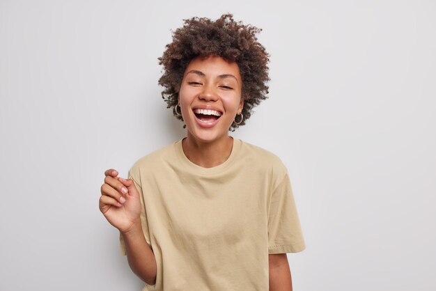 긍정적인 곱슬머리의 아름다운 여성은 흰색 스튜디오 배경에 격리된 캐주얼한 베이지색 티셔츠를 입고 평온한 표정을 유지하며 행복하게 웃고 있습니다. 진실한 인간의 감정 개념