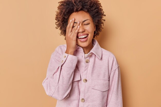Positive curly haired brunette woman smiles positively makes face palm keeps eyes closed dressed in pink jacket isolated over beige background. Sincere dark skinned female model feels joyful