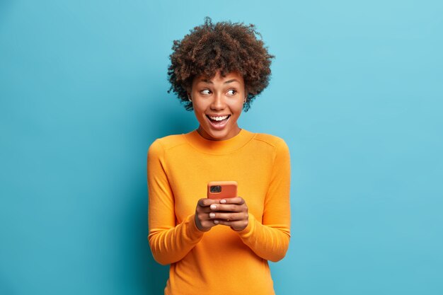 Positive curly haired ethnic woman uses mobile phone checks messages and reads news holds modern cellular in hands looks with curious happy expression on right isolated over blue wall