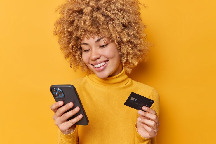 Free Photo positive curly haired european woman smiles happily uses modern smartphone and credit card purchases something makes shopping online dressed in casual jumper isolated over vivid yellow background
