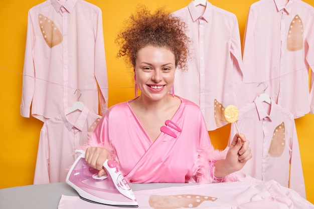 ポジティブな巻き毛のヨーロッパの女性は、洗濯室の黄色い壁に隔離されたピンクのガウンを着てロリポップを着て服をなでるためにスチームアイロンを使用しています。アイロンと家事のコンセプト