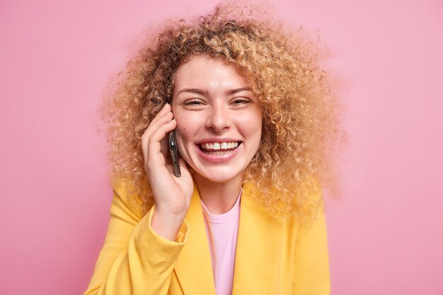 Positive curly haired female model has happy look enjoys pleasant telephone talk smiles toothily shows teeth dressed in yellow jacket isolated over pink wall chuckles during conversation