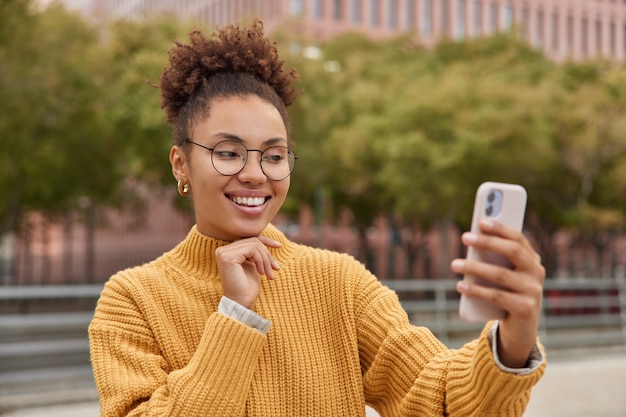 Positive curly haired girl creats selfie content via smartphone spends leisure time uses mobile technology for shooting vlog wears big round spectacles and yellow knitted sweater stands outdoors