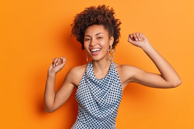 Positive curly haired lady dances with arms raised smiles broadly wears fashionable polka dot blouse isolated over vivid orange wall has happy mood