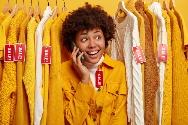 販売中の流行の服を購入して幸せなポジティブな巻き毛の女性、衣類ラックに立って、