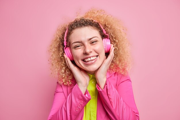Позитивная кудрявая девочка-подросток любит слушать любимую музыку в стереонаушниках, в хорошем настроении носит яркую одежду, изолированную от розовой стены. Счастливый женский меломан слушает песню