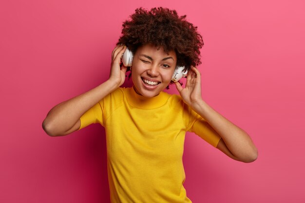 Positive curly haired teenager winks eye, smiles happily as listens music in stereo headphones, feels amused, tilts head, enjoys nice sound, wears yellow t shirt, poses against pink wall