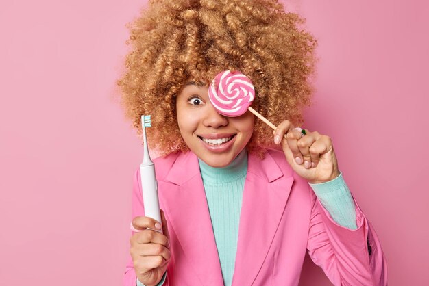 Positive curly haired woman covers eye with delicious candy holds electric toothbrush has sweet tooth dressed in elegant clothes isolated over pink background Teeth problem and harmful food