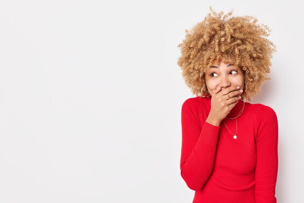 Positive curly haired woman covers mouth giggles positively tries to hide emotions laughs at something funny dressed in red poloneck isolated over white background blank copy space for your text