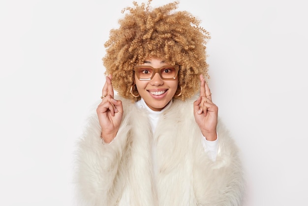 Positive curly haired woman dressed in winter fur coat crosses fingers believes in good luck hopes dreams come true smiles pleasantly wears transparent spectacles isolated over white background