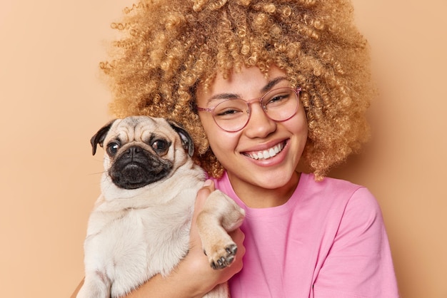 긍정적인 곱슬머리 여성은 좋아하는 퍼그 개를 껴안고 갈색 배경에서 격리된 시력 교정을 위해 행복한 안경을 쓰고 있는 애완동물과 즐거운 시간을 보냅니다. 우정과 가축 개념