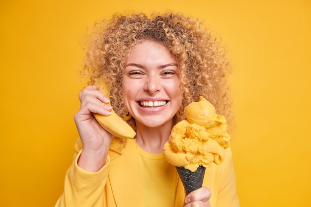 Positive curly haired woman has fun enjoys eating delicious ice cream of lemon flavor keeps banana near ear pretends calling someone expresses positive emotions isolated over yellow wall