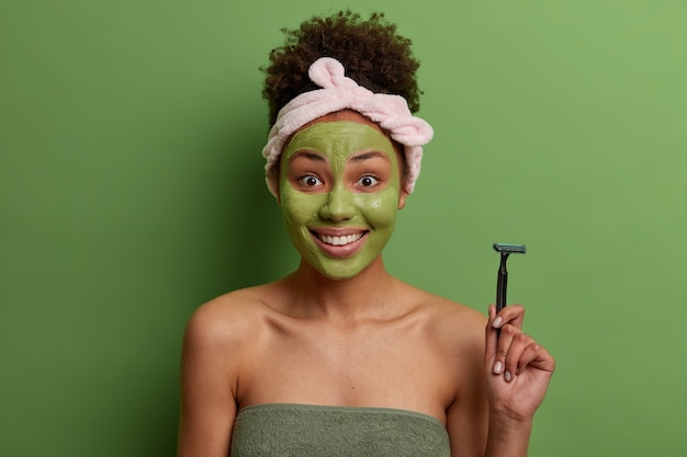 긍정적 인 곱슬 머리 여자는 면도기를 들고 다리를 면도하고 얼굴에 보습 마스크를 바르고 목욕 타월에 싸서 녹색 벽 위에 절연되어 있습니다. 웰빙, 순결, 위생