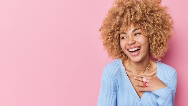 Foto gratuita la giovane donna dai capelli ricci positiva distoglie lo sguardo con un'espressione gioiosa sorride ampiamente esprime emozioni felici indossa un maglione blu casual isolato su sfondo rosa copia spazio per la tua promozione