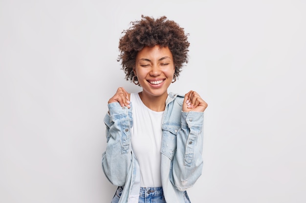 Positive curly haired young woman raises hands awaits for results rejoices awesome news keeps eyes closed smiles broadly wears denim shirt isolated over white wall