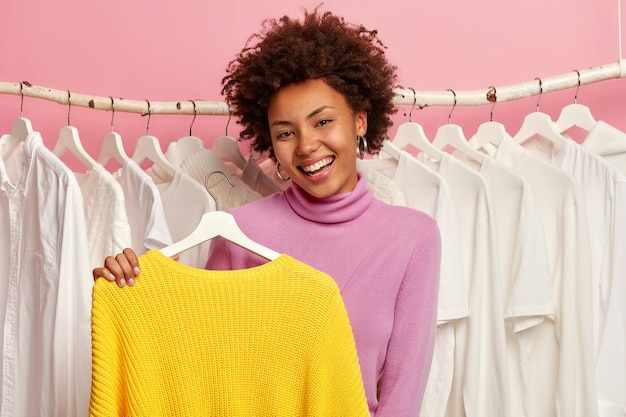 Positive curly woman holds yellow jumper on hangers, tries fresh new apparel, selects outfit for casual occasion, has happy expression