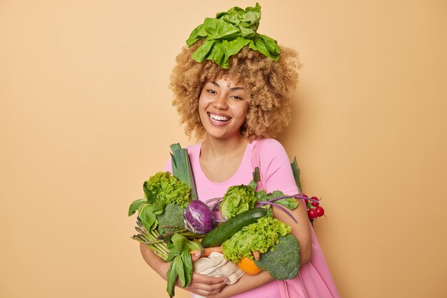 Позитивная кудрявая женщина позирует со свежими зелеными овощами, полными витаминов, рада богатому урожаю, радостно улыбается, носит повседневную футболку, изолированную на коричневом фоне Концепция здорового питания