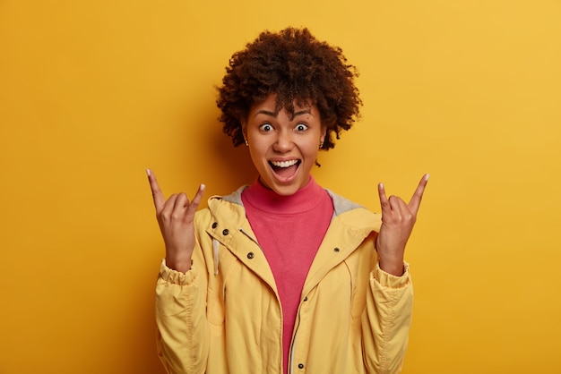 Free photo positive curly woman vibes at awesome concert, enjoys party, makes rock on gesture, heavy metal sign, being fan of rock songs, dressed in jacket, isolated on yellow wall, feels carefree