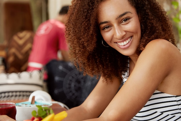 Positive curly young woman with dark healthy skin, smiles pleasantly, sits at cafe table surrounded with delicious dish, enjoys recreation time in restaurant. People, lifestyle and ethnicity concept