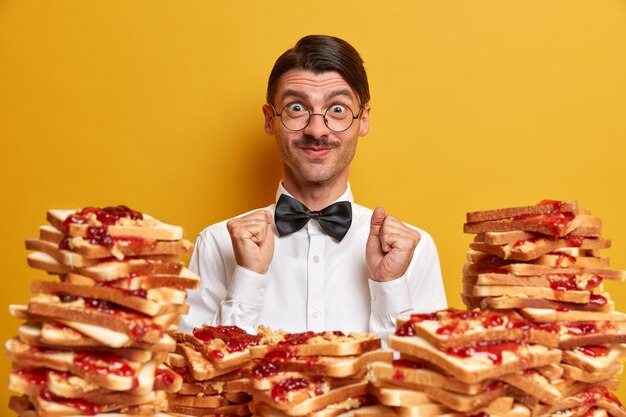 Positive customer rejoices all inclusive in restaurant, stands near pile of delicious toasts, wears elegant white shirt and bowtie, clenches fists, 