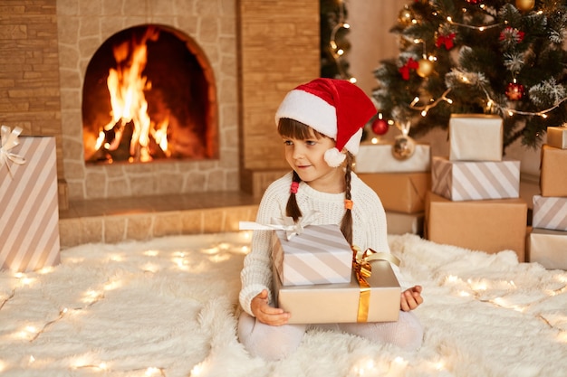 Positive cute little girl wearing white sweater and santa claus hat, holding stack of presents in hands, sitting on floor on soft carpet near Christmas tree, present boxes and fireplace.