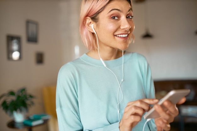 Positive cute student girl having fun indoors after college using headset while chatting online via video chat on her smartphone