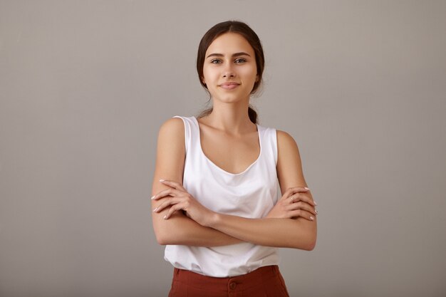 Positive cute young European woman crossing arms confidently on her chest and smiling joyfully posing in empty apartment with freshly painted blank walls with copyspace for you advertising content