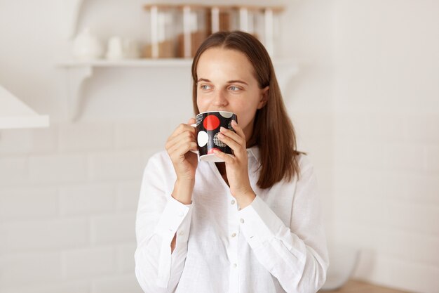 Позитивная темноволосая женщина в белой футболке повседневного стиля, пьет кофе или чай по утрам, позирует с кухонным гарнитуром, смотрит в сторону с мечтательным выражением лица.