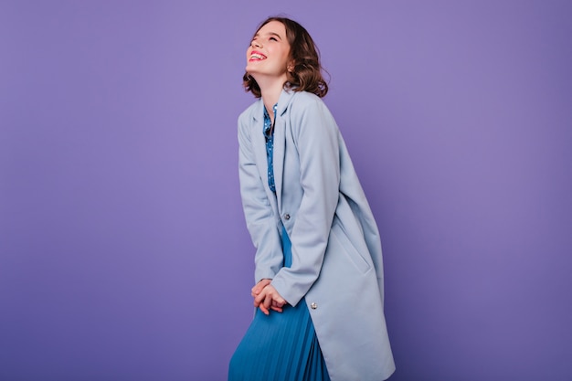 Positive dark-haired girl smiling during photoshoot in blue coat. amazing curly woman expressing positive emotions on purple wall.