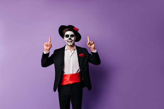 Positive dark-haired guy in Mexican-style clothes joyfully shows his fingers upwards, posing for portrait with space for text.