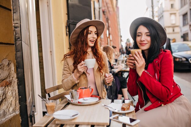 Позитивная темноволосая женщина в красной куртке пьет кофе с закрытыми глазами со своей подругой