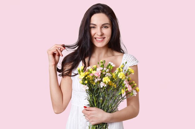 positive dark haired woman touches hair, smiles gently, has appealing look, holds first spring flowers, has red manicure, dressed in white dress, isolated on rosy. Beauty concept