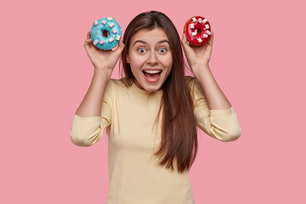 Positive dark haired young woman holds two sweet doughnuts, keeps mouth widely opened