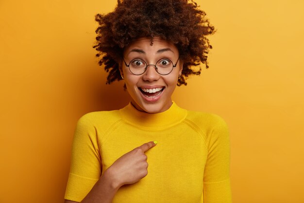 Positive dark skinned Afro American woman points at herself, asks who me, happy to be chosen or to win, wears bright yellow clothes, poses indoor. Happy reaction, good emotions and feelings.