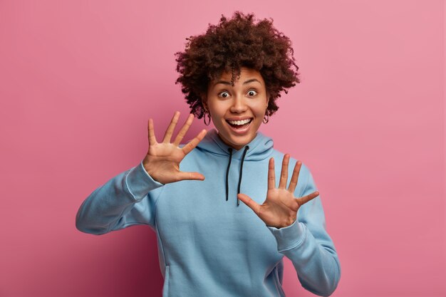 Positive dark skinned Afro American woman raises palms, laughs happily, has widely opened eyes, glad reaction, playful mood, giggles positively, wears blue sweatshirt, isolated on pink wall.