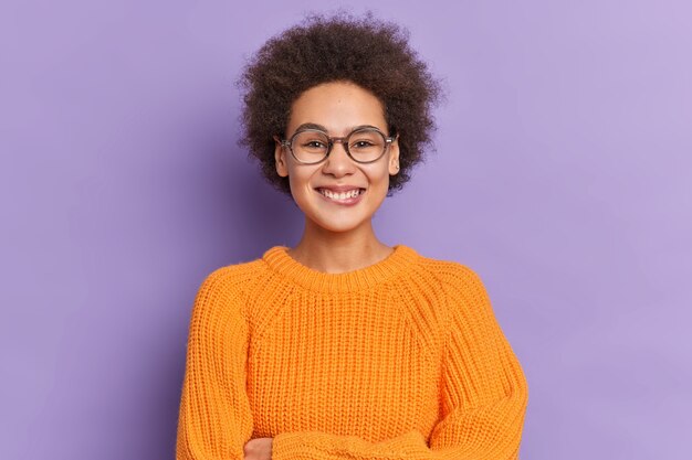 Positive dark skinned beautiful teenage girl with bushy Afro hair smiles happily dressed in knitted orange sweater and spectacles.