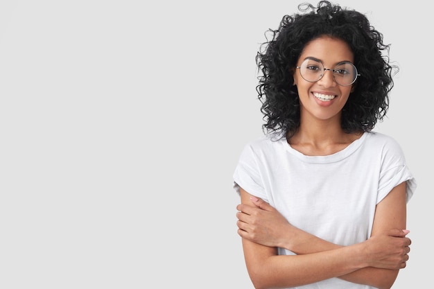 La femmina dalla pelle scura positiva ha un sorriso a trentadue denti, un'espressione timida, tiene le braccia conserte, sembra felice, felice di parlare con un amico, isolato sul muro bianco