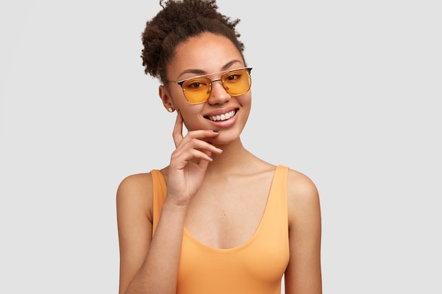 Positive dark skinned female model in trendy sunglasses, casual vest, has white perfect teeth, stands over wall, expresses happiness. Happy young African American woman models indoor