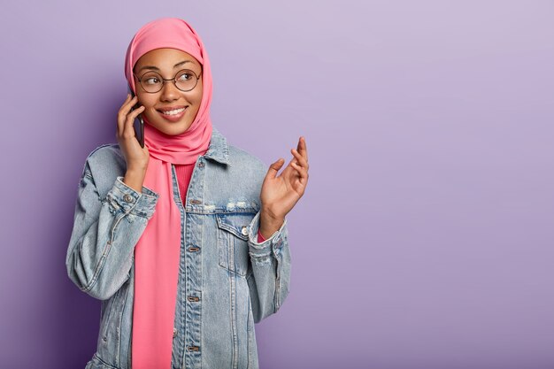 Positive dark skinned female wears traditional Muslim scarf on head, keeps cell phone near ear, enjoys pleasant conversation, gestures while tells something exciting to interlocutor isolated on purple