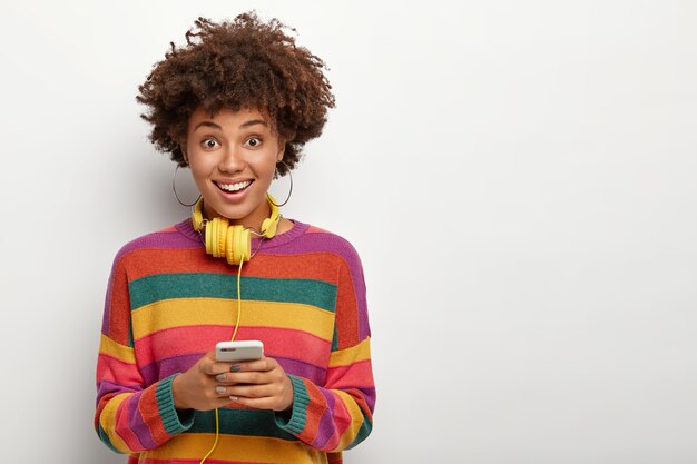 Positive dark skinned girl checks audio quality with headphones, listens music, wears striped sweater, poses over white background.